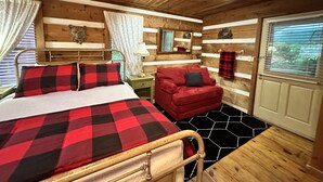 A wooden cabin bedroom with a bed, a red sofa, a nightstand, and a window with blinds. - Queen Cozy Cabin on Main St (Blowing Rock)