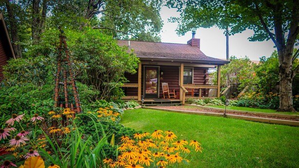 A rustic log cabin with a porch, surrounded by a well-maintained garden with yellow flowers and green foliage.