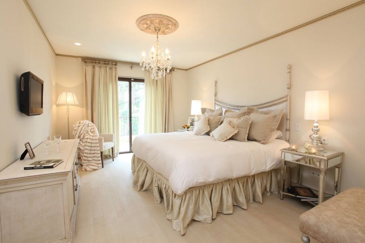 A bedroom with a large bed, a bedside table, a desk, a chair, a TV, a chandelier, and a window with curtains.