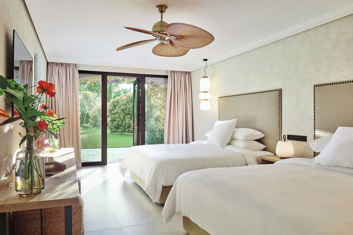 A hotel room with two beds, a ceiling fan, a bedside table with a lamp, and a view of a garden through the window.