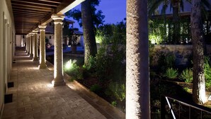 A covered walkway with stone columns and a brick floor, surrounded by greenery and palm trees. - Hotel Cigarral de las Mercedes (Toledo)