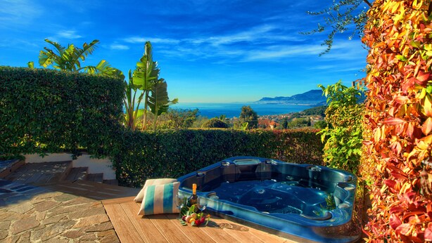 A hot tub with a wooden deck and a bottle of wine, surrounded by lush greenery and a view of the ocean. - Two Bedroom Apartment with Pool, Jacuzzi, A/C, WiFi and Electric Bicycles (Bordighera)