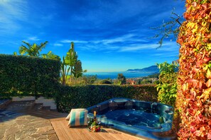 A hot tub with a wooden deck and a bottle of wine, surrounded by lush greenery and a view of the ocean. - Two Bedroom Apartment with Pool, Jacuzzi, A/C, WiFi and Electric Bicycles (Bordighera)