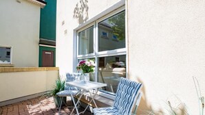 Eine Terrasse im Freien mit Tisch und gestreiften Stühlen, einem Blumentopf und einem Fenster mit Blick auf ein Gebäude.