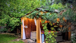 A wooden gazebo with a bench and a hot tub, surrounded by greenery and stone pathways.