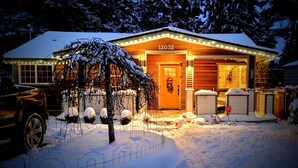 A wooden cabin with a number 12032, surrounded by snow, with a porch and a vehicle parked nearby.
