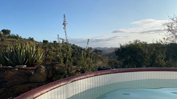 Une piscine bordée d’un mur en pierre, avec des plantes d’un côté, et une vue sur des collines et des arbres en arrière-plan.