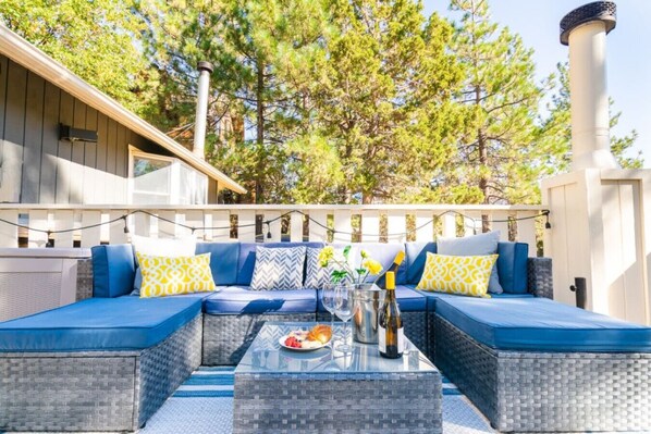 An outdoor seating area with a blue sofa, yellow patterned pillows, and a glass table with a bottle and glasses.