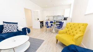 Uma sala de estar moderna com um sofá azul, uma poltrona amarela e uma mesa de centro branca. Há uma área de jantar com cadeiras azuis e uma mesa de vidro. A cozinha é visível ao fundo, com armários brancos e bancada preta.