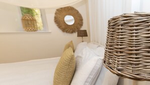 Cama com roupa de cama branca e duas almofadas bege, candeeiro de vime, espelho redondo na parede e uma pequena janela com vista para vegetação.