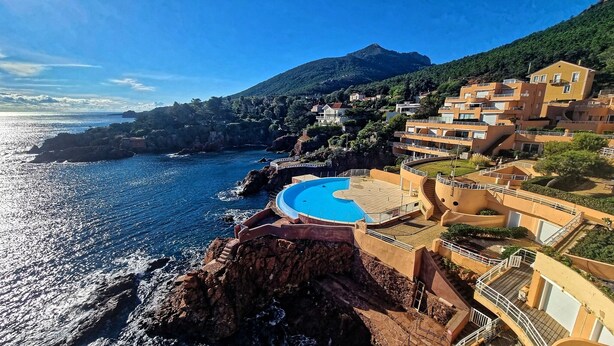 Un hotel frente al mar con piscina, balcones escalonados y montañas al fondo.