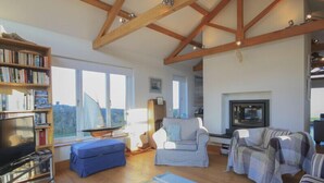 A spacious living room with a high ceiling, a fireplace, a bookshelf, and a large window.