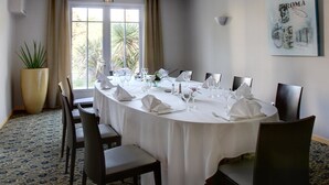 Un salón de conferencias con una larga mesa dispuesta para una reunión, ventanas amplias con cortinas y una planta en maceta.