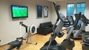 Un gimnasio en casa con bicicletas estáticas, una cinta de correr y un televisor mostrando un partido de fútbol.