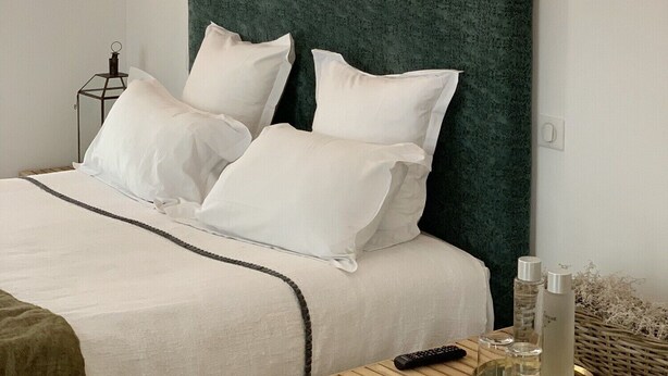 Una cama bien tendida con sábanas blancas y un cabecero verde, una mesita de noche con un control remoto, botellas y un cuaderno.