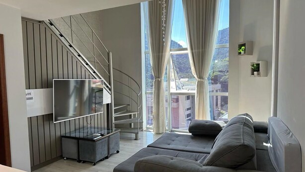 Een moderne woonkamer met een grijze bank, een flatscreen televisie aan de wand, een trap met een metalen leuning en een groot raam met gordijnen.