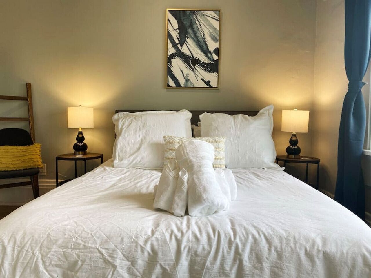 A neatly made bed with white linens and towels, two bedside lamps, and a painting on the wall.