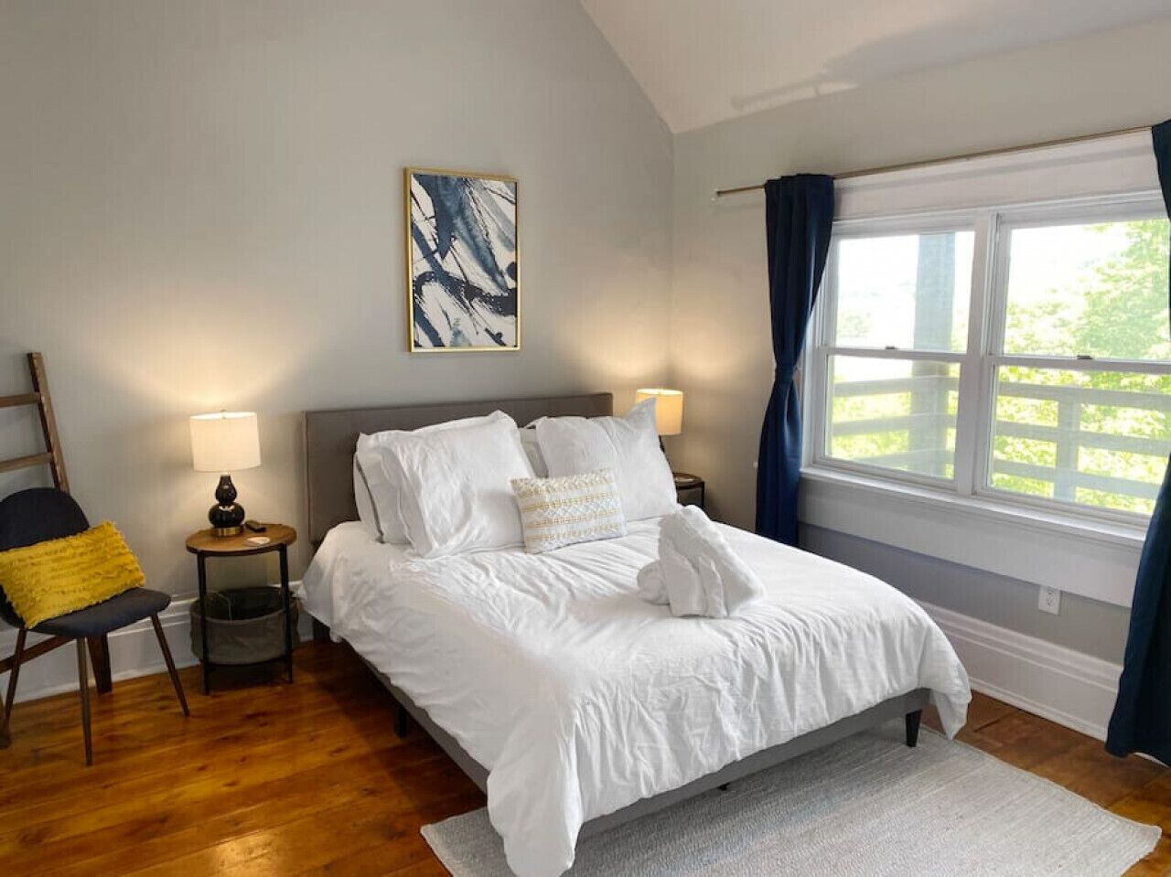 Een slaapkamer met een bed, twee nachtkastjes, een stoel, een raam met gordijnen en een schilderij aan de muur.