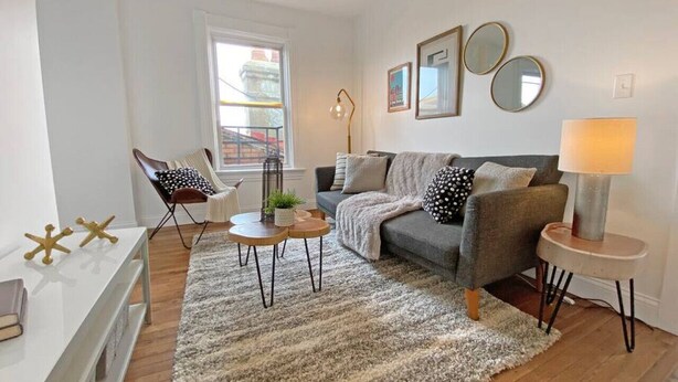 A living room with a grey sofa, a white coffee table, a small side table, and a lamp. There is a window with a view of buildings outside. - Explore Downtown and Mount Adams with River Views! (Cincinnati)