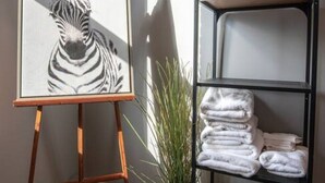 Quarto com uma pintura de zebra emoldurada em um cavalete, uma planta em vaso, uma estante de metal com toalhas dobradas e uma janela projetando uma sombra.