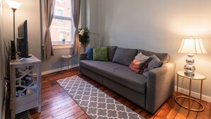 Uma sala de estar com um sofá cinzento, uma mesinha lateral, um abajur e uma TV sobre um suporte.