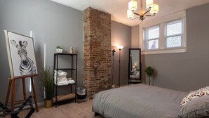 Quarto com parede de tijolos, uma cama, um quadro em cavalete, uma prateleira com toalhas dobradas, um espelho e uma janela com persianas.