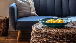 A modern living room with a dark blue sofa, a wicker ottoman, and a framed artwork on the wall.