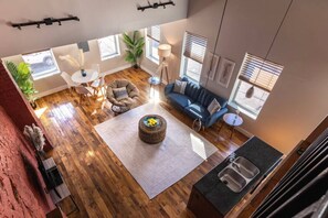 A spacious living room with a large window, a blue sofa, a round coffee table, and a kitchen area with a sink. - Broadway Suites - Stunning Downtown Condos (Cincinnati)