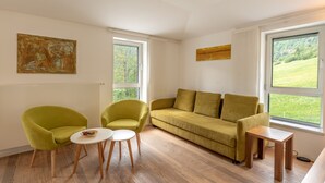 A modern living room with a sofa, two armchairs, a coffee table, and a small side table. There are large windows offering a view of greenery outside. - Apartment Glacier View 14.6. with balcony (Kaprun)