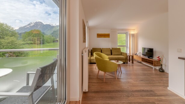 A modern living room with a sofa, yellow armchair, and a TV on a wooden console. A glass door leads to a balcony with a mountain view. - Apartment Glacier View 14.6. with balcony (Kaprun)
