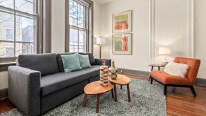 Un salon moderne avec un canapé gris, une table basse en bois et un fauteuil rouge. Il y a deux fenêtres avec des stores, une lampe et des œuvres d’art encadrées accrochées au mur.