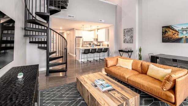 A modern living room with a spiral staircase, a leather sofa, a wooden coffee table, and a kitchen in the background. - Soaring 2-Bedroom Parkside Loft Walkable to it All (Cincinnati)