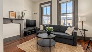 A modern living room with a sofa, a coffee table, a TV, and large windows. - Deluxe 5-bedroom Condo Walkable to Everything (Cincinnati)