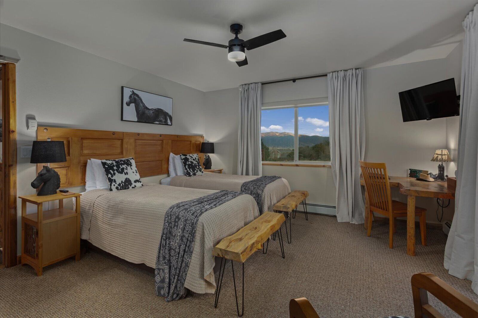 Habitación de hotel con dos camas, un escritorio, una silla, un televisor y ventana con vista a la montaña.