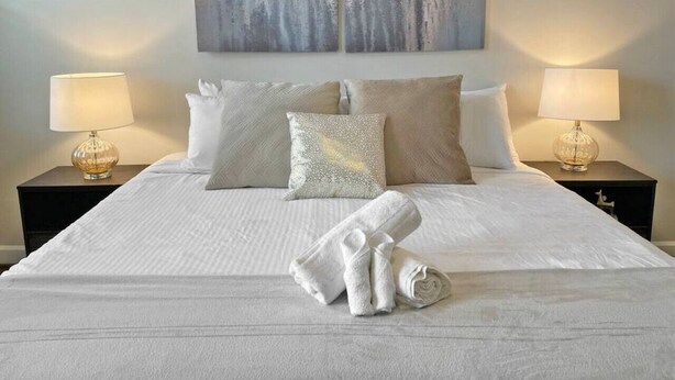 Een netjes opgemaakt bed met witte lakens, twee kussens en een opgevouwen handdoek. Twee nachtkastjes met lampen en een aan de muur hangend kunstwerk boven het bed.