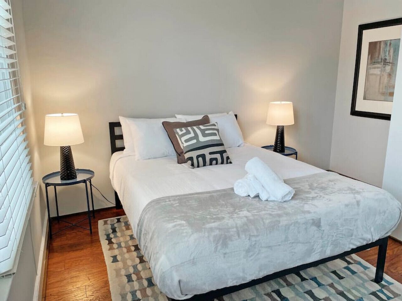 A neatly made bed with a grey duvet, two pillows, and a folded towel on a patterned rug.