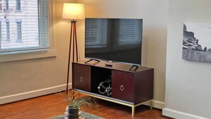 Een woonkamer met een flatscreen-televisie op een donkere houten tv-meubel, een vloerlamp en een decoratieve plant op een glazen tafel.