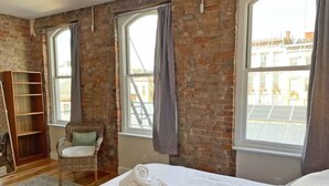 A hotel room with a large bed, a wicker chair, a wooden bookshelf, and a view of buildings through large windows. - Findlay Suites - Perfect Location Over the Rhine (Cincinnati)