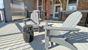 Un espacio exterior techado con sillas Adirondack y una mesa redonda, al lado de un edificio con fachada de ladrillo y ventanas grandes.