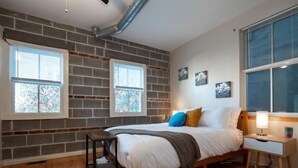 Ein Schlafzimmer mit Backsteinwand, Holzboden, einem Bett mit Kissen, einem Nachttisch und einem Fenster mit Jalousien.