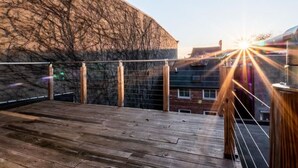 A wooden deck with a metal railing, overlooking a building with a textured facade. - Cozy OTR Home with Private Roof Deck (Cincinnati)