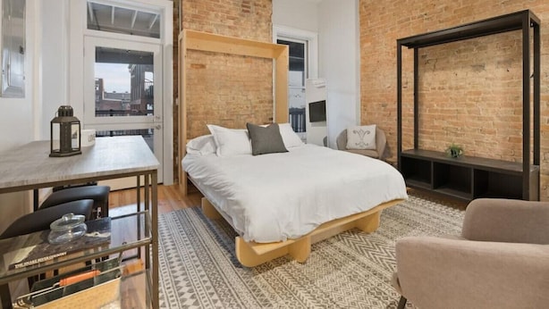 A bedroom with a bed, a desk, a chair, a TV, and a brick wall. - Over the Rhine Luxury MicroSuite On Findlay Market (Cincinnati)
