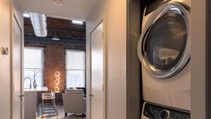 A laundry room with two stacked front-loading washers and dryers, a kitchen area with a sink and bar stools, and a brick accent wall.
