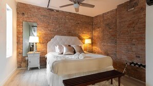 A bedroom with a brick wall, a bed with white bedding, a nightstand with a lamp, and a bench.