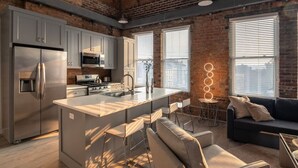 A modern kitchen with a large island, stainless steel appliances, and a brick wall. There is a seating area with a sofa and chairs.