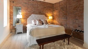 A bedroom with a brick wall, a tufted headboard, a wooden bench, and a nightstand with a lamp. - Entire Historic Building: Sleeps 30 (Cincinnati)