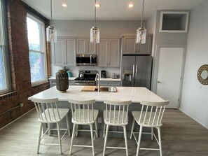 Una cocina moderna con armarios blancos, una isla central con taburetes de bar, electrodomésticos de acero inoxidable e iluminación colgante.