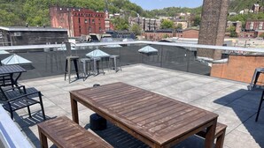A rooftop patio with a wooden table and benches, surrounded by umbrellas and a view of the cityscape.