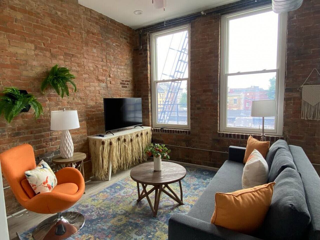 Un salón con pared de ladrillo, un ventanal grande, un sofá, un sillón naranja, un televisor y una mesita redonda.