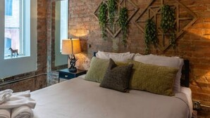 Un dormitorio con pared de ladrillo, cama con ropa de cama blanca, ventilador de techo y ventana con vistas.